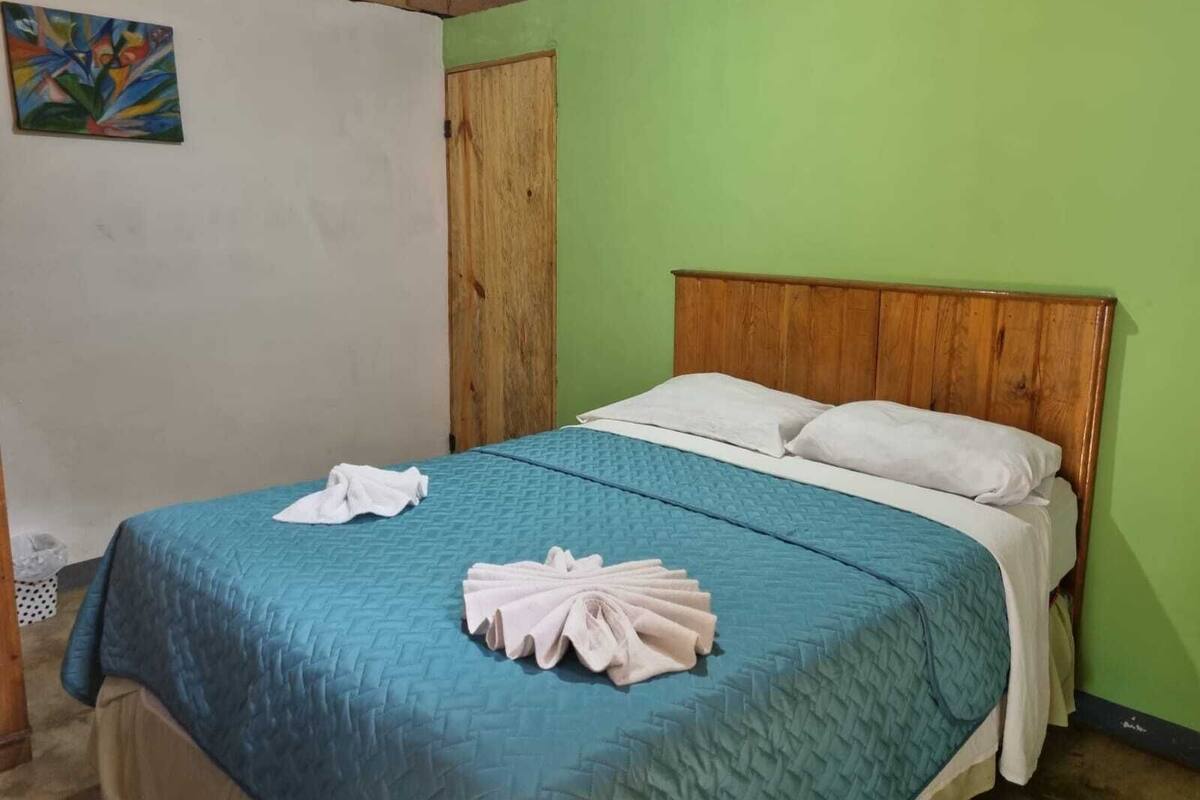 A single bed with a wooden headboard, a blue quilt, and white pillows. There is a towel folded on the bed and a painting on the wall.