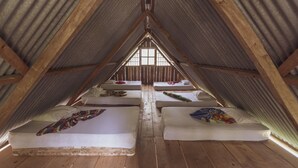 Een zolderkamer met vier bedden gerangschikt in een U-vorm, houten balken en een groot raam aan het einde van de kamer.