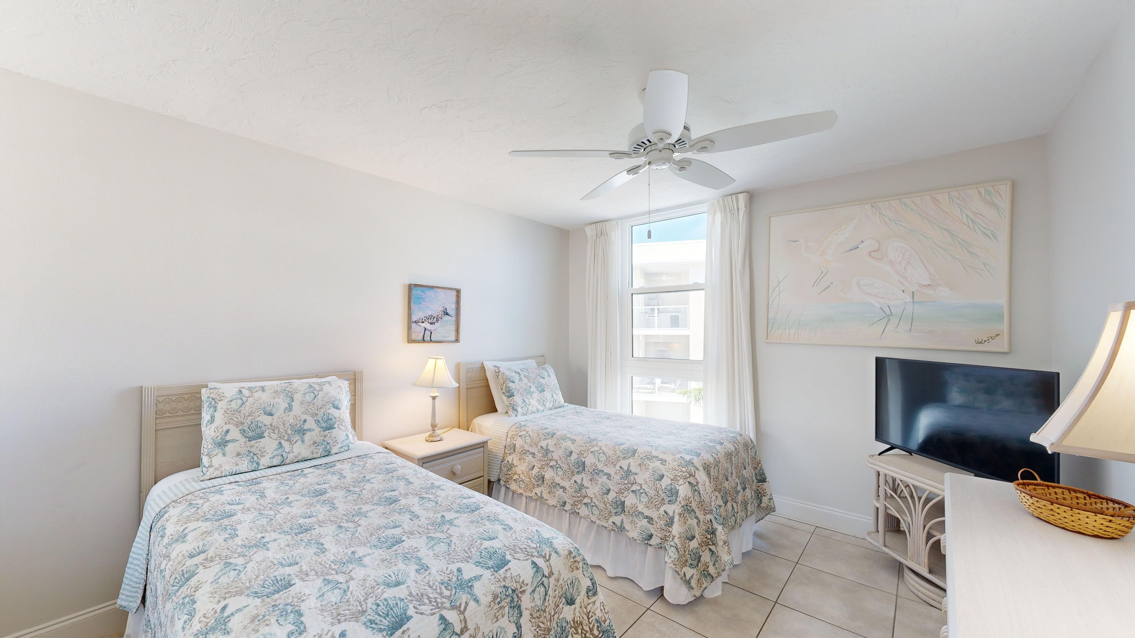 A hotel room with two beds, a ceiling fan, a TV, and artwork on the walls.