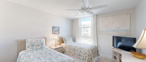 A hotel room with two beds, a ceiling fan, a TV, and artwork on the walls.