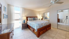 A bedroom with a bed, bedside tables, a ceiling fan, a large mirror, and a view of the outside.