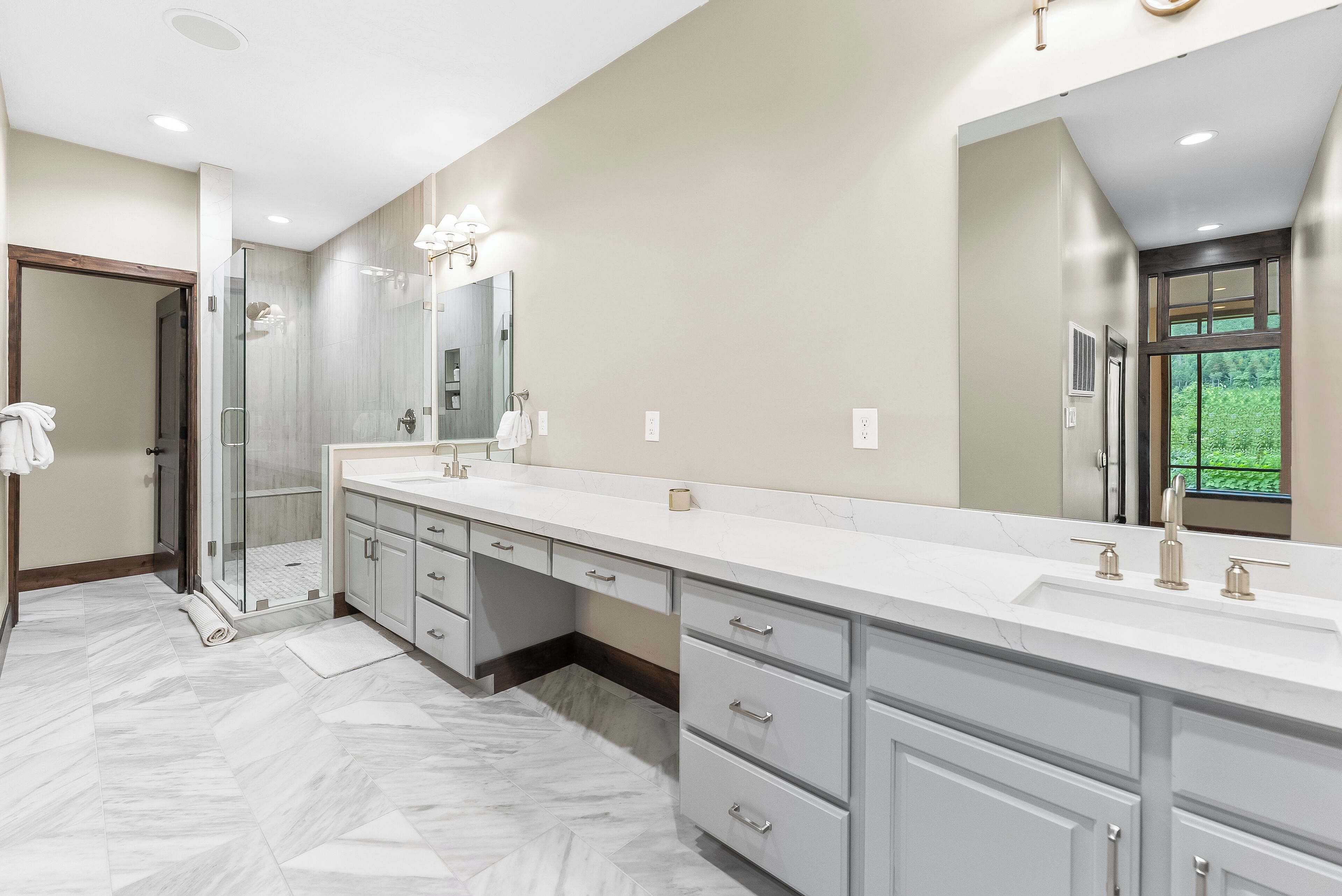 A modern bathroom with a large vanity, a glass shower enclosure, and a view of greenery outside.