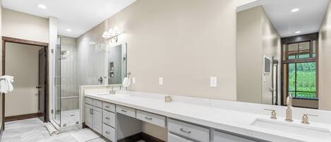 A modern bathroom with a large vanity, a glass shower enclosure, and a view of greenery outside.