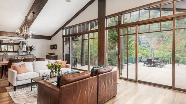 A spacious living room with a large window, a ceiling fan, and a dining area visible through the window. - Mountain Lodge Overlooking Waterfall - Hot Tub, 5 bedroom, Unbelievable Views! (Sundance)