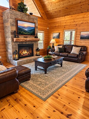 A log cabin interior with a stone fireplace, leather furniture, and a large window.