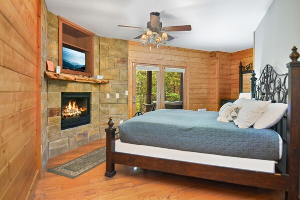 A bedroom with a stone fireplace, a bed with a blue quilt, a ceiling fan, and a TV mounted on the wall.