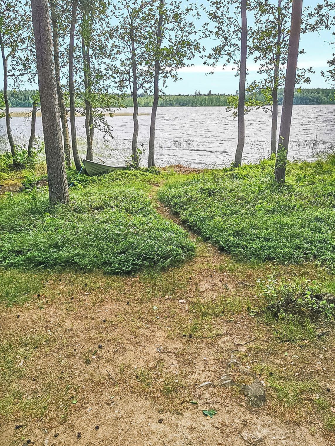 Luonnonmaisema, jossa on maaperäinen polku, joka johtaa vesistöön. Polun ympärillä on puita ja vehreää kasvillisuutta.