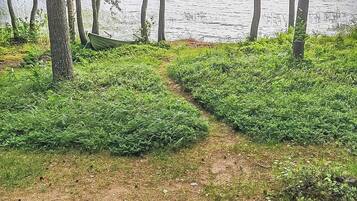 Luonnonmaisema, jossa on maaperäinen polku, joka johtaa vesistöön. Polun ympärillä on puita ja vehreää kasvillisuutta.