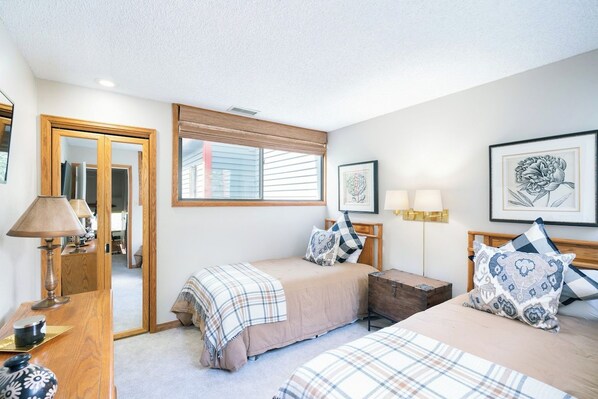 A bedroom with two beds, a bedside table, a lamp, a mirror, and a window with blinds.