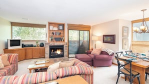 A living room with a fireplace, a television, a dining area with a table set for two, and a large window with a view of trees. - Telluride Lodge #532 2 Bedroom Condo (Telluride)