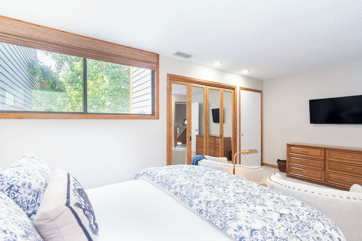A bedroom with a large window, a bed with patterned bedding, a dresser, and a television mounted on the wall.