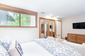 Une chambre avec une grande fenêtre, un lit avec une literie à motifs, une commode et un téléviseur fixé au mur.