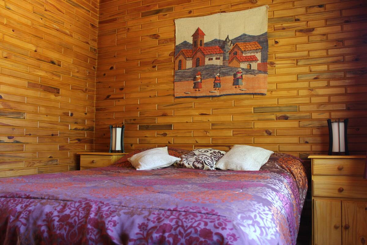 Una habitación con paneles de madera, una cama con colcha morada estampada, dos mesitas de noche y un tapiz que muestra una escena de pueblo.