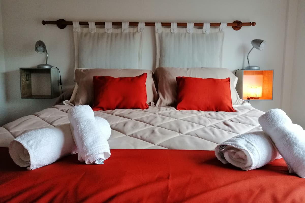 Una cama bien hecha con una colcha roja y beige, dos almohadas rojas y dos toallas blancas enrolladas.