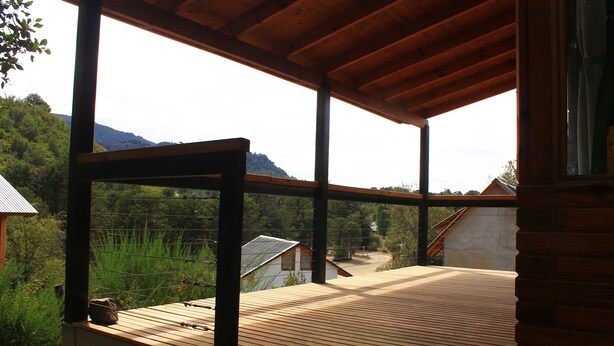 Una terraza de madera con vistas a un paisaje rural, que incluye una casa y montañas.