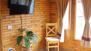 Interior de una cabaña de madera con un televisor en la pared, una planta en maceta, una silla de madera, un sofá morado y una ventana con cortinas.