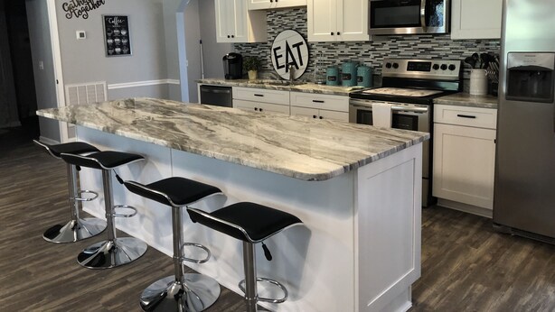 Una cucina moderna con mobili bianchi, un'isola in marmo con sgabelli da bar, elettrodomestici in acciaio inossidabile e un cartello con la scritta 'Cater Together'.