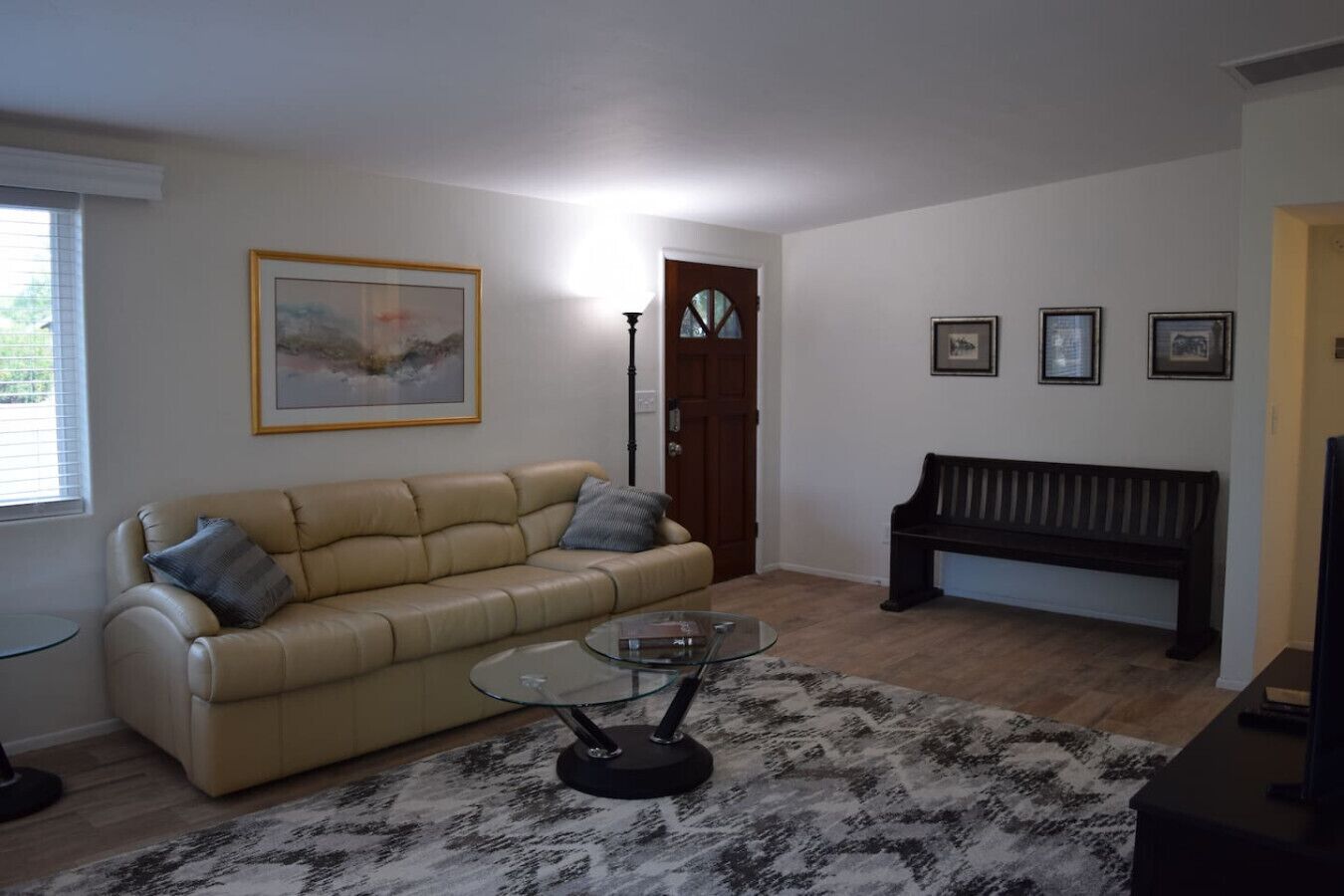 A living room with a beige sofa, a glass coffee table, a black bench, and framed pictures on the wall.