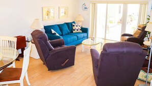 Un salon avec un canapé bleu, deux fauteuils violets, une table basse en verre et une table à manger blanche avec des chaises.