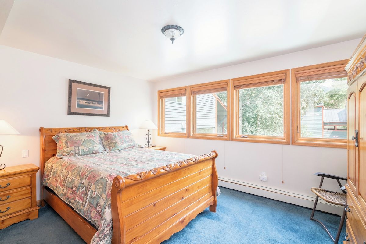 A bedroom with a wooden bed, a nightstand, a lamp, a chair, and a window with a view of trees.