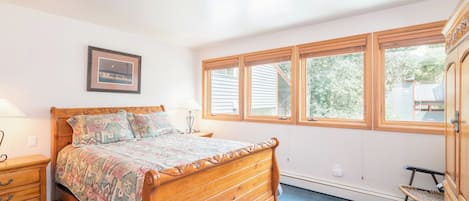 A bedroom with a wooden bed, a nightstand, a lamp, a chair, and a window with a view of trees.