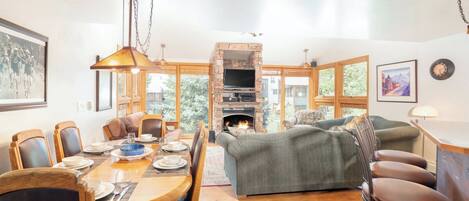 A dining area with a wooden table and chairs, a fireplace, and a sofa in a bright room with large windows.