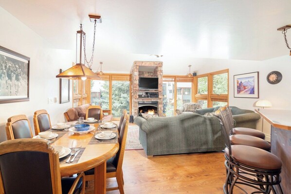 A dining area with a wooden table and chairs, a fireplace, and a sofa in a bright room with large windows.