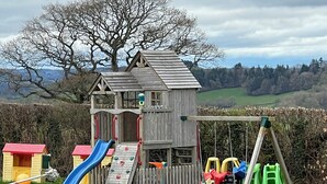 A wooden playhouse with a slide and climbing wall, surrounded by a swing set and picnic table. - Visit Wales 4 Star, rural bungalow - Sauna, jacuzzi, play area and Free Wi-fi  (Pontypool near Usk, Panteg)