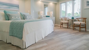 A bedroom with a bed, bedside tables, a chair, and a window with blinds. - G-101 Port O’ Call - “Ocean Breeze” - with Ocean Views - Pool Access. (Isle of Palms)