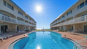 A swimming pool with a sun shining above, surrounded by a building with balconies and lounge chairs.