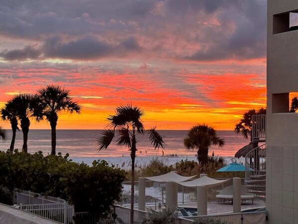 A sunset view from a balcony overlooking the ocean with palm trees and a building structure. - House Of The Sun #212GV (Sarasota)