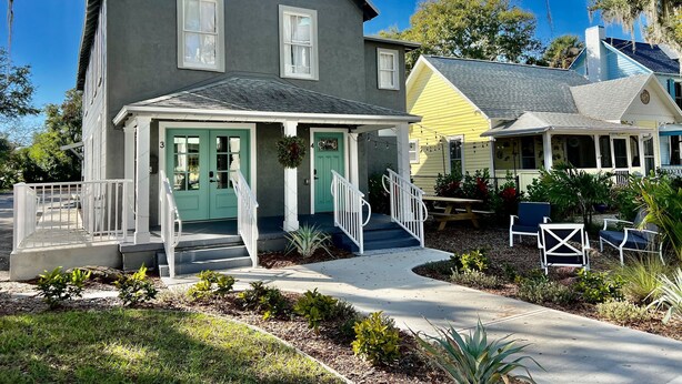 A two-story house with a porch, a front door, and a small front yard with plants and a bench.