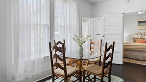 A dining area with a round glass table surrounded by wooden chairs, a vase with flowers, and sheer white curtains.