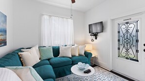 A teal-colored sofa with multiple cushions, a white coffee table with a remote control, a patterned rug, a ceiling fan, a TV mounted on the wall, and a door with a 'No Smoking' sign.