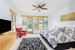 Une chambre à coucher avec un lit, un téléviseur, un fauteuil rouge, un ventilateur de plafond et de grandes fenêtres.