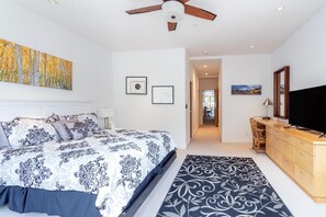A bedroom with a large bed, a ceiling fan, a TV, and a desk.