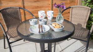A round glass table with a metal frame, surrounded by wicker chairs. On the table, there are plates, bowls, a vase with flowers, and a mug.