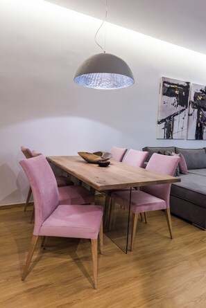 Un coin repas avec une table en bois et des chaises recouvertes d’un tissu rose, un luminaire suspendu gris et, au fond, un canapé gris.