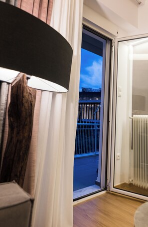 A room with a wooden floor, a white radiator, a sliding glass door leading to an outdoor balcony with a metal railing, and a lamp with a white shade. - Acropolis Panoramic View Deluxe Apartment | 3 Falirou str 11742 Athens (Athina)