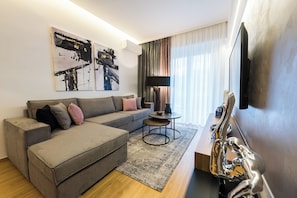 A modern living room with a grey sectional sofa, a small round table, and a flat-screen TV mounted on the wall. - Acropolis Panoramic View Deluxe Apartment | 3 Falirou str 11742 Athens (Athina)