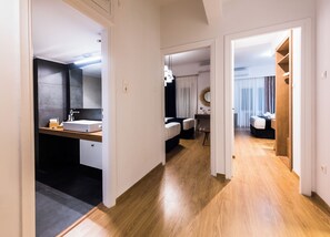 A hotel room with a wooden floor, a bathroom with a dark tile wall, a bed with a white headboard, and a desk with a mirror. - Acropolis Panoramic View Deluxe Apartment | 3 Falirou str 11742 Athens (Athina)
