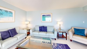 A living room with a glass coffee table, two sofas with cushions, a side table with a lamp, and two framed pictures on the wall.