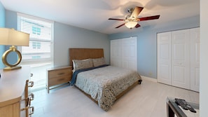 A bedroom with a bed, nightstand, lamp, ceiling fan, and a desk with a lamp.