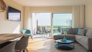 Uma sala de estar moderna com uma mesa de jantar de madeira, um sofá e uma mesa de centro. Há uma porta de vidro deslizante que dá para uma área externa com vista para a paisagem.