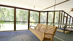 Eine Holzbank in einem überdachten Bereich mit großen Fenstern, einem Deckenventilator und Blick auf Bäume.
