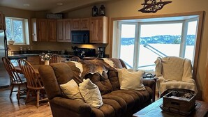 A cozy living room with a brown sofa, a wooden coffee table, and a view of a snowy landscape through large windows. - Welcome to our Mountain Lake Getaway (White Sulphur Springs)