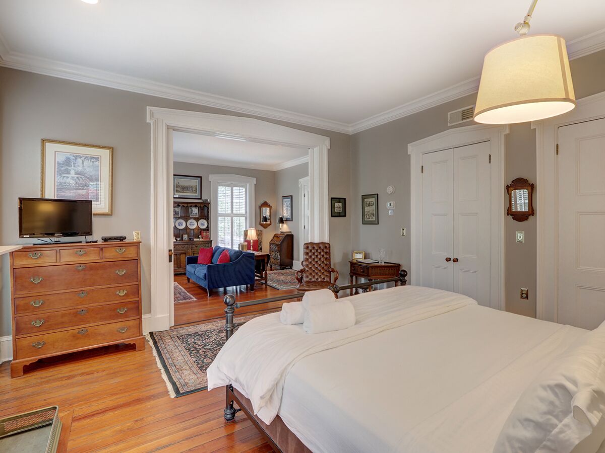 A bedroom with a large bed, wooden dresser, and a television on a dresser. There is a hallway leading to another room with a blue sofa and a chair.