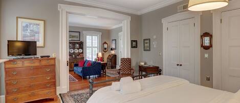 A bedroom with a large bed, wooden dresser, and a television on a dresser. There is a hallway leading to another room with a blue sofa and a chair.