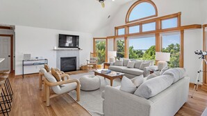A spacious living room with a fireplace, large windows, and a ceiling fan. - Sound Decision: Hot Tub | Sound Views | Pool Access (Nags Head)