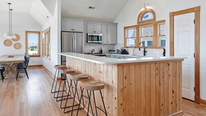 A modern kitchen with a large island, wooden bar stools, and a dining area with a table and chairs. - Sound Decision: Hot Tub | Sound Views | Pool Access (Nags Head)